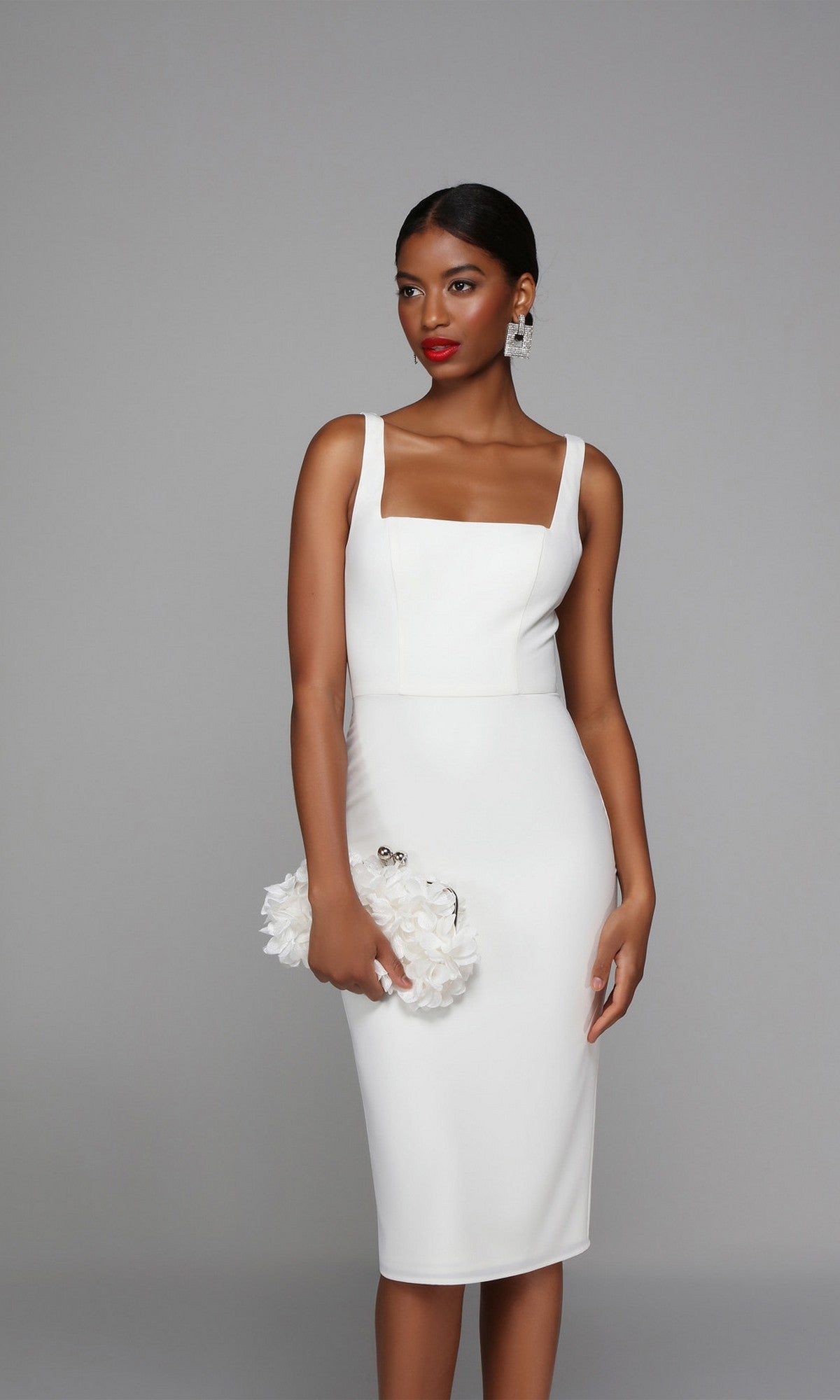 Woman wearing the knee length semi-formal Alyce 70010 dress in ivory, holding a white floral clutch, standing against a plain gray background.